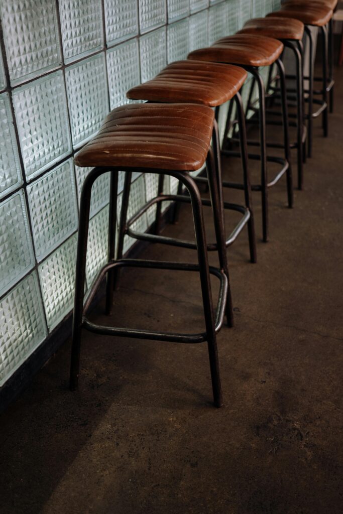 pexels-photo-5530255-5530255 Row of leather and metal bar stools against a textured glass wall, showcasing modern industrial design.